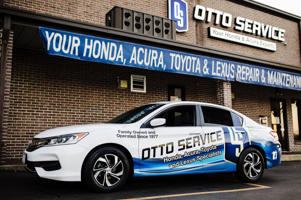 A branded Otto Service car is parked in front of a building with a banner reading "Your Honda, Acura, Toyota & Lexus Experts.